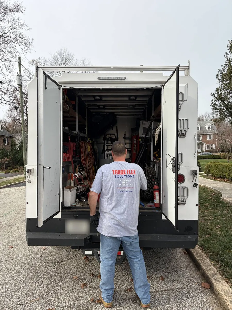 Plumbarama technician in a Trade Flex Solutions shirt pulling equipment from the back of a service truck on a residential street